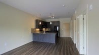 This single-floor space offers a thoughtfully designed open-concept layout that seamlessly connects the kitchen and living areas. The kitchen is a standout feature, showcasing dark espresso-toned cabinetry that extends to the upper walls, providing generous storage while creating a bold, contemporary aesthetic. A large center island with a dark wood-panel base and a stone-look countertop serves as both a functional workspace and a natural gathering point. Elegant pendant lights hang above the island, casting a warm, focused glow that complements the under-cabinet tile backsplash in a neutral subway pattern. Black stainless steel appliances, including a refrigerator and range with microwave, add a sleek, cohesive finish to the culinary space. Rich, dark hardwood-style flooring flows throughout the entire unit, grounding the space with warmth and continuity. The open living area is bathed in soft, ambient light from recessed ceiling fixtures, contributing to an inviting and well-lit atmosphere. Smooth white ceilings with clean lines enhance the sense of openness. A hallway visible in the background suggests additional private spaces beyond the main living area, completing this well-appointed, modern residence.