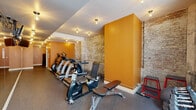 This fitness center in New Orleans, LA, blends industrial character with modern workout amenities in a thoughtfully designed single-level space. The room is defined by striking exposed brick walls with a weathered, textural finish that lend a raw, historic aesthetic, contrasted by bold amber-orange accent walls and structural columns that add warmth and visual interest throughout.

The cardio zone is well-stocked, featuring multiple treadmills, elliptical trainers, and a stationary bike arranged along the length of the space, allowing for comfortable circulation between machines. A large floor-to-ceiling mirror along one wall visually expands the room and is practical for monitoring form during workouts. The free weights area is situated near a generous grid-pane window that allows natural light to fill the corner, and includes a tiered dumbbell rack, a adjustable weight bench, a medicine ball, and plyometric equipment.

Rubber flooring throughout provides durability and comfort underfoot. Ceiling-mounted globe light fixtures provide consistent, ambient illumination across the space. A restroom with exposed brick walls and tile flooring is accessible directly from the fitness area, adding convenience for users. The overall ambiance is energetic yet grounded, balancing New Orleans' architectural heritage with functional fitness infrastructure.
