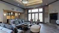 This well-appointed single-floor space exudes a refined, contemporary elegance designed for comfort and social gathering. The room's most prominent architectural feature is its coffered tray ceiling, highlighted by a central flush-mount light fixture that draws the eye upward and adds a sense of height and grandeur to the interior.

Large French-style doors and expansive windows line the far wall, allowing an abundance of natural light to flood the space while offering clear views of the surrounding greenery outdoors. Sheer and panel curtains frame the windows, adding softness to the otherwise structured design.

The seating arrangement is thoughtfully curated, featuring a generously sized grey sofa adorned with patterned throw pillows, complemented by upholstered accent chairs in coordinating tones. A dark wood coffee table anchors the main seating area, while a large round ottoman in a neutral fabric adds a versatile, contemporary touch.

To one side, a bar-height counter with pendant lighting hints at a refreshment or service area. Decorative wall plates and framed artwork contribute subtle character throughout. The flooring transitions between plush area rugs and hard surface tile, enhancing both comfort and visual interest across the space.