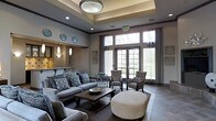 This well-appointed single-floor space exudes a refined, contemporary elegance designed for comfort and social gathering. The room's most prominent architectural feature is its coffered tray ceiling, highlighted by a central flush-mount light fixture that draws the eye upward and adds a sense of height and grandeur to the interior.

Large French-style doors and expansive windows line the far wall, allowing an abundance of natural light to flood the space while offering clear views of the surrounding greenery outdoors. Sheer and panel curtains frame the windows, adding softness to the otherwise structured design.

The seating arrangement is thoughtfully curated, featuring a generously sized grey sofa adorned with patterned throw pillows, complemented by upholstered accent chairs in coordinating tones. A dark wood coffee table anchors the main seating area, while a large round ottoman in a neutral fabric adds a versatile, contemporary touch.

To one side, a bar-height counter with pendant lighting hints at a refreshment or service area. Decorative wall plates and framed artwork contribute subtle character throughout. The flooring transitions between plush area rugs and hard surface tile, enhancing both comfort and visual interest across the space.