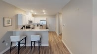 This single-floor unit offers a thoughtfully designed layout with a clean, contemporary aesthetic and functional living spaces throughout. The kitchen serves as a central highlight, featuring crisp white shaker cabinetry, granite countertops with a subtle speckled pattern, a subway tile backsplash, and a full suite of stainless steel appliances including a range, over-the-range microwave, and French door refrigerator. A peninsula with seating provides a casual dining option and connects seamlessly to the open living and dining area, which is finished with warm wood-look flooring and neutral walls that create a welcoming atmosphere. The living area accommodates comfortable seating and features window blinds that allow for natural light control. Two bedrooms offer carpeted floors and a calm, restful ambiance, each with access to natural light. A spacious walk-in closet with wire shelving provides generous storage. The unit includes two full bathrooms, each with white vanity cabinetry, integrated sinks, and tile flooring. One bathroom features a shower-tub combination, while the other offers a clean, open layout. Recessed lighting and consistent neutral tones tie the entire space together cohesively.