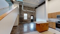 Located at 601 W 2nd St in Davenport, Iowa, this two-story loft blends industrial character with modern finishes across a well-appointed layout. The expansive main floor is anchored by soaring ceilings with exposed ductwork and structural beams, original brick accent walls, and polished concrete floors that reflect natural light streaming in through large windows. A staircase leads to the upper level, with additional storage tucked conveniently beneath it. The kitchen is a standout feature, offering warm wood cabinetry, granite countertops, stainless steel appliances including a range, microwave, refrigerator, and dishwasher, and a central island ideal for dining or meal prep. Both bathrooms are finished with matching granite vanities and warm wood cabinetry, complemented by terrazzo-style tile flooring. The primary bedroom provides a comfortable retreat with a built-in closet and terrazzo flooring. The upper floor offers a more intimate mezzanine-style space. Throughout the home, the interplay of raw industrial elements and refined finishes creates a distinctive urban loft ambiance that is both functional and full of character.
