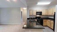 This single-floor 1-bedroom, 1-bathroom residence offers a thoughtfully designed open-concept layout that seamlessly connects the kitchen and living areas. The kitchen serves as a central focal point, showcasing dark granite countertops with subtle speckling that contrast elegantly against the light wood cabinetry above. A peninsula with an integrated sink extends into the space, providing additional prep area and defining the boundary between the kitchen and living area. Black appliances, including an electric range and over-the-range microwave, complement the dark countertop aesthetic. The cabinetry offers ample storage with a clean, natural wood finish that keeps the space feeling bright and open. Neutral-toned walls throughout the unit create a cohesive, versatile backdrop, while soft carpeting in the living area adds warmth underfoot. A hallway visible from the main living space leads to the private bedroom and bathroom, ensuring a practical and logical flow throughout the unit. Recessed and flush-mount ceiling lighting provides consistent illumination across the space. Overall, the unit balances functional design with modern finishes in a compact, efficient layout.