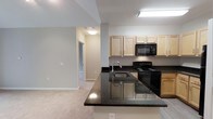This single-floor 1-bedroom, 1-bathroom residence offers a thoughtfully designed open-concept layout that seamlessly connects the kitchen and living areas. The kitchen serves as a central focal point, showcasing dark granite countertops with subtle speckling that contrast elegantly against the light wood cabinetry above. A peninsula with an integrated sink extends into the space, providing additional prep area and defining the boundary between the kitchen and living area. Black appliances, including an electric range and over-the-range microwave, complement the dark countertop aesthetic. The cabinetry offers ample storage with a clean, natural wood finish that keeps the space feeling bright and open. Neutral-toned walls throughout the unit create a cohesive, versatile backdrop, while soft carpeting in the living area adds warmth underfoot. A hallway visible from the main living space leads to the private bedroom and bathroom, ensuring a practical and logical flow throughout the unit. Recessed and flush-mount ceiling lighting provides consistent illumination across the space. Overall, the unit balances functional design with modern finishes in a compact, efficient layout.