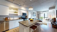 This contemporary single-floor urban residence showcases an industrial-modern aesthetic defined by striking exposed concrete ceilings throughout. The open-concept layout seamlessly connects the kitchen and living areas, creating a cohesive and functional living space bathed in natural light from oversized windows.

The kitchen is well-appointed with white cabinetry, a mosaic tile backsplash, stainless steel appliances including a refrigerator, electric range, and microwave, and a central island with a sleek countertop and wooden bar stools — ideal for casual dining or entertaining. Track lighting and a pendant fixture complete the space.

The living area features warm hardwood flooring, large floor-to-ceiling windows that invite ample natural light and urban views, and direct access to a private balcony. The exposed concrete ceiling carries throughout, reinforcing the space's industrial character.

The bedroom is a tranquil retreat with plush carpeting, a concrete ceiling, and a generous window offering views of the surrounding cityscape. Two bathrooms serve the unit: one features a tiled tub-shower combination with warm wood-look flooring, while the second offers a glass-enclosed walk-in shower, an extended double vanity, and large-format tile flooring. Together, these spaces deliver a refined, urban living experience.