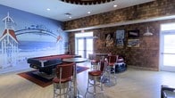 This single-floor recreational lounge is designed around a lively retro boardwalk theme, delivering a nostalgic atmosphere that is both engaging and visually dynamic. The space is anchored by a bold coastal mural depicting a classic seaside amusement park, rendered in vivid blues and warm tones that immediately draw the eye upon entry. Rustic cedar shake-style wall cladding adds texture and warmth throughout, evoking the charm of a vintage beach boardwalk.

Seating is arranged around round bistro-style tables paired with chrome and red vinyl bar stools — a nod to mid-century American diner aesthetics. A pool table occupies one corner, offering a casual recreation option alongside the lounge seating. Decorative touches include retro amusement-themed posters and signage mounted along the walls, reinforcing the playful boardwalk narrative. A novelty bumper car repurposed as a seating piece adds a whimsical, interactive element to the décor.

Recessed lighting illuminates the space from a stylized ceiling featuring a scalloped border detail, while natural light filters in through glass entry doors. The light wood flooring complements the warm, earthy wall tones, tying the overall design together. The result is an inviting, entertainment-focused environment full of character and retro charm.