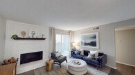 This single-floor residence offers a thoughtfully designed layout that balances comfort and functionality across its key living areas. The living room serves as the heart of the home, anchored by a striking white-painted brick fireplace with a dark wood mantle. Rich hardwood-style flooring, sliding glass doors, and vertical blinds fill the space with natural light and provide access to an outdoor balcony. The kitchen features white shaker-style cabinetry, a marble-look tile backsplash, stainless steel appliances including a full-size refrigerator, and a breakfast bar that connects seamlessly to the rest of the living area. Adjacent to the kitchen, the dining area accommodates a round table for four beneath a modern chandelier, with a sliding glass door that further extends the natural light throughout. Both bedrooms are finished with plush carpeting, neutral-toned walls, and generous windows dressed with curtain panels, creating restful and inviting retreats. The bathroom features wood-look tile flooring, gray vanity cabinetry with ample storage, a single basin sink, and a separate tub and shower area. Throughout the residence, textured ceilings and a cohesive neutral palette provide a clean, comfortable backdrop suited to a variety of lifestyles.