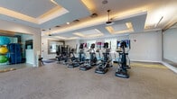 Located at 1 John F Kennedy Blvd in Somerset, NJ, this fitness center offers a comprehensive workout environment spread across a single thoughtfully designed floor. The main exercise area is spacious and open, accommodating an extensive row of modern cardio machines — including ellipticals, stair climbers, and treadmills — each equipped with individual display screens for an engaging workout experience. Pendant ceiling fans are suspended from elegant tray ceilings with recessed lighting, lending the space a polished, upscale aesthetic. Large mirrors line portions of the walls, enhancing the sense of openness throughout the room. A dedicated strength training zone features a multi-station cable machine, free weights, and additional resistance equipment, all set on durable, patterned carpet tile flooring with yellow accent strips. A glass-walled spin studio provides a semi-private space for cycling workouts, with a row of stationary bikes visible within. Natural light filters in through multiple windows, creating a bright and inviting atmosphere. Exterior-facing doors offer easy access and further contribute to the airy feel of the space. The overall design balances functionality with a clean, modern aesthetic, making this fitness center a well-appointed destination for a full range of exercise activities.