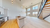 This contemporary two-floor residence is defined by its dramatic double-height ceilings and open, airy layout. The main living area is anchored by a sleek kitchen island topped with dark granite countertops, a brushed gold sink fixture, and pendant lighting that hangs from the soaring ceiling above. White cabinetry with brushed metal hardware lines the kitchen walls, complemented by a subtle glass tile backsplash and stainless steel appliances. Light hardwood flooring flows throughout, enhancing the sense of openness.

A striking staircase with steel treads and a cable railing system connects the two floors, with a stacked washer and dryer tucked neatly beneath it. Large black-framed windows along the main living space flood the interior with natural light and frame urban views of the surrounding cityscape. Upper-floor rooms continue the light hardwood flooring and feature additional windows that capture expansive outdoor views.

The bathrooms are finished to a high standard, featuring large-format marble-look tile flooring, granite vanity tops with white cabinetry, and frameless glass-enclosed walk-in showers with neutral tile surrounds. Throughout the space, the design is cohesive and modern, with a neutral color palette, clean lines, and thoughtful finishes that create a bright, sophisticated living environment.
