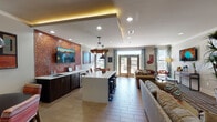 This single-floor communal lounge is designed with an open-concept layout that seamlessly blends a kitchenette, dining, and multiple lounge areas into one cohesive, inviting space. The kitchenette serves as a central hub, featuring a large white island with bar-height seating, dark cabinetry with a built-in sink, under-counter refrigeration, and a wall-mounted flat-screen TV set against a striking textured brick accent wall. Pendant lighting — including industrial black fixtures and a rustic wood chandelier — adds warmth and character throughout. Adjacent to the kitchenette, a casual dining nook with a round table and colorful striped chairs sits beneath a pendant light, accented by eclectic artwork on the walls. The lounge area is generously furnished with plush sofas, leather accent chairs, a striped area rug, and a wall-mounted television, creating a relaxed and social atmosphere. Large glass French doors flood the space with natural light and provide direct access to an outdoor area. Wood-look tile flooring flows throughout, tying the zones together. The overall design balances industrial and eclectic aesthetics, making it a versatile and welcoming gathering space.
