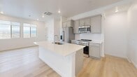 Located on W Wrigley Dr in Lehi, Utah, this contemporary three-story home offers three bedrooms, three bathrooms, and a thoughtfully organized layout spread across three distinct floors. The ground level houses a generously sized attached garage with an automatic opener, concrete flooring, and direct interior access — providing ample parking and storage space. The main living floor features a bright, open-concept area combining the kitchen, dining, and living spaces. The kitchen is appointed with gray shaker-style cabinetry, a white quartz waterfall island with an integrated sink, stainless steel appliances including a gas range and French door refrigerator, and a classic white subway tile backsplash. Light hardwood flooring runs throughout this level, complementing the clean, neutral palette. The upper floor hosts all three bedrooms, including the primary suite with light hardwood floors, multiple windows framing expansive Utah desert and mountain views, and a sliding door to the balcony. Additional bedrooms feature plush carpet and corner windows that flood rooms with natural light and scenic vistas. The bathrooms are finished with tile flooring, sliding glass shower enclosures, and clean white vanities. A dedicated laundry area with stacked units and four closets — including a walk-in — add practical storage throughout.
