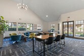 Located at 1256 West Arrow Highway in Upland, CA, this single-floor community clubhouse offers a generously sized, thoughtfully designed gathering space. The living room is the heart of the space, boasting dramatic vaulted ceilings, striking sputnik chandeliers, and large grid-pane windows that flood the interior with natural light and offer views of the surrounding greenery. A bold teal lattice-panel feature wall anchors a media area furnished with plush seating, while a built-in shelving unit with a flat-screen TV provides an additional lounge setting. Colorful patterned carpet tiles in blue and grey tones run throughout, adding a contemporary, artistic touch. A dedicated business center features a sleek marble-topped workstation with multiple monitors, complemented by high-top tables set against wide windows. The game room area includes a ping pong table beneath vaulted ceilings with direct access to the outdoors through glass double doors. The kitchen is well-appointed with light wood cabinetry, white quartz countertops, stainless steel appliances including a range, microwave, dishwasher, and refrigerator, and warm hardwood-style flooring. A dining area with a long communal table rounds out this vibrant, multifunctional clubhouse.