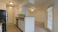 This single-floor residence offers a thoughtfully designed open-concept layout that connects the kitchen and living area in a natural, flowing arrangement. The kitchen is well-appointed with white cabinetry providing generous storage space, granite-look countertops, a stainless steel dishwasher, and a built-in sink. A peninsula-style breakfast bar extends from a structural column, creating a convenient divide between the kitchen and the adjoining living space while keeping the areas visually connected. The flooring transitions from tile in the kitchen to carpeting in the living area, adding warmth and definition to each zone. The living area is spacious and open, bathed in a bright, neutral palette that enhances the sense of airiness throughout. A ceiling fan with a light fixture is centrally positioned in the living space, providing both air circulation and ambient lighting. The clean white walls and understated finishes throughout lend the space a fresh, versatile character well-suited for a variety of interior styles. The overall layout is practical and efficient, making excellent use of the available floor plan while maintaining an inviting and comfortable atmosphere.