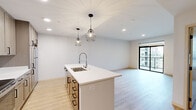 This thoughtfully designed single-floor residence blends modern aesthetics with functional living. The open-concept kitchen and living area serves as the heart of the home, anchored by a sleek central island with a white quartz countertop, an integrated stainless steel sink, and stylish geometric cage pendant lights overhead. The kitchen is equipped with stainless steel appliances, including a range, microwave, and refrigerator, complemented by warm taupe cabinetry, white subway tile backsplash, and recessed lighting. Light wood-tone flooring flows seamlessly throughout the entire space, enhancing the bright, airy atmosphere created by crisp white walls.

The bedrooms are generously proportioned and filled with natural light from large black-framed windows and sliding glass doors that open to private balconies overlooking the surrounding urban landscape. One bedroom features a stylish barn door leading to a walk-in closet with built-in shelving. The bathroom is clean and well-appointed, with a light wood vanity, white countertop, and a shower-tub combination. Additional bedrooms offer grid-pane windows that invite natural light while maintaining a modern aesthetic. The overall ambiance is fresh, minimalist, and move-in ready.