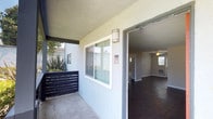 This single-floor unit offers a straightforward and functional layout designed for comfortable everyday living. The entry is accessed via a covered outdoor walkway, providing a semi-private transition space before stepping inside. A wall-mounted exterior light fixture illuminates the entryway, and a slatted privacy screen adds a touch of separation from the surrounding environment, complemented by low-maintenance desert-style plantings nearby.

The interior is defined by rich, dark hardwood-style flooring that runs throughout the visible living areas, lending warmth and a sense of cohesion to the space. Neutral gray walls provide a clean, versatile backdrop that suits a variety of furnishing styles. Sliding windows along the exterior wall allow for natural light to filter into the unit, while a ceiling fan is visible deeper within, supporting airflow and comfort.

The open sightlines from the entryway into the main living area convey a sense of openness, with additional rooms visible toward the rear of the unit. The textured exterior stucco finish and shared staircase nearby are characteristic of a well-organized multi-unit residential building. Overall, the space presents a clean, move-in-ready environment with classic, enduring finishes throughout.