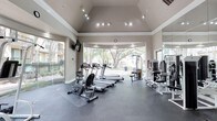 This fitness center offers a bright and functional environment designed to support a well-rounded workout routine. The space is defined by its striking vaulted ceiling, which lends an open, airy feel and is complemented by track lighting that ensures even illumination throughout. Large floor-to-ceiling windows along the back wall invite an abundance of natural light while offering views of lush, mature trees outside, creating a refreshing and motivating atmosphere. The flooring consists of durable, dark rubberized material well-suited for high-activity use. Cardio equipment, including treadmills, ellipticals, and a rowing machine, is arranged thoughtfully to make the most of the available floor space. A dedicated strength training area features a cable machine and additional resistance equipment, while full-length mirrors along one wall help users maintain proper form and visually expand the room. A wall-mounted television provides entertainment during workouts. A glass door offers easy access to the outdoor surroundings, further enhancing the connection to the natural environment. The overall design balances practicality with an inviting aesthetic, making this fitness center a comfortable and well-appointed space for daily exercise.