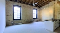 This single-floor space showcases a distinctive industrial-loft character, defined by its exposed yellow brick walls and open wood beam ceiling construction. The raw, structural ceiling reveals original wooden joists and is equipped with a ceiling fan, contributing to both the aesthetic and practical functionality of the space. A track lighting system runs along one wall, offering flexible illumination options suited for a variety of uses.

Large, black-framed windows with arched detailing line the main wall, allowing generous amounts of natural light to flood the interior while providing views of the surrounding urban environment. The windows are complemented by baseboard heating units positioned beneath them, ensuring comfort throughout the seasons.

The flooring features a smooth, light-colored concrete or epoxy surface that contrasts warmly with the textured brick and wood elements above. The open, unobstructed floor plan provides a highly versatile layout adaptable to creative, commercial, or studio purposes. The combination of historic architectural details with functional modern fixtures gives this space a compelling and characterful ambiance.