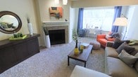 This thoughtfully designed single-floor space offers a comfortable and welcoming atmosphere throughout. The living area serves as the heart of the home, centered around a classic fireplace framed with mosaic tile surround and a clean white mantel, above which decorative artwork adds a pop of color and personality. Soft, neutral carpeting extends across the living area, complementing the warm tones of the walls and creating a cozy underfoot feel. A large window dressed with blue curtains floods the space with natural light, brightening the open layout and offering views of the surrounding greenery. The living room is well-appointed with a mix of modern and casual furnishings, including a wood coffee table, a bold orange accent chair, and a textured pouf, all contributing to the space's relaxed yet stylish character. Adjacent to the living area, a compact dining nook with a bistro-style table and chairs takes advantage of the natural light from the windows, making it an ideal spot for casual meals. A dark wood console adds contrast and anchors one side of the room, providing additional storage and display space. Overall, the space strikes a balance between functional design and comfortable living.