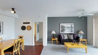 This single-floor residence showcases a thoughtfully designed interior that balances contemporary style with everyday functionality. The living area serves as the heart of the space, anchored by a striking slate-grey accent wall that provides a dramatic backdrop for wall art and furnishings. The room is furnished with a dark upholstered sofa accented with grey and purple throw pillows, complemented by warm yellow wood furniture pieces, including a coffee table and side tables, creating a vibrant contrast against the neutral palette. A framed cityscape print adds an urban, sophisticated touch to the living space. The flooring transitions seamlessly from rich hardwood in the entry and hallway areas to plush carpeting in the living room, defining each zone with warmth and texture. Decorative wall clocks and framed artwork along the hallway walls add personality and depth to the space. A hallway visible from the living area leads to additional rooms, hinting at a well-connected floor plan. The clean white ceilings and walls throughout keep the space feeling bright and open, while the carefully curated décor adds layers of visual interest. Overall, this residence offers a modern, welcoming atmosphere with a distinct design sensibility.