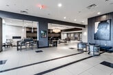 This expansive lobby presents a polished, contemporary design characterized by large-format cream tile flooring accented with dark inlay strips that guide movement throughout the space. The open layout is organized into several distinct functional zones, each contributing to the overall sophistication of the environment.

At the entrance, floor-to-ceiling glass walls and black-framed partition doors flood the space with natural light and provide a transparent, welcoming threshold. Adjacent to the entry, a comfortable seating area features mid-century modern-style chairs, an upholstered ottoman, and a round area rug centered around a wooden accent table, creating an inviting gathering point.

The primary lounge area anchors the space with a bold navy blue sofa, coordinating accent chairs, low coffee tables, and textured ottomans arranged beneath a dramatic drum chandelier. A built-in mailbox wall spans the background, adding a practical yet architectural element. A dedicated dining area, separated by deep navy accent walls and pendant lighting—including a cascading crystal chandelier—offers seating around sleek dark tables. A linear fireplace enhances the ambiance of this zone.

The elevator corridor features stainless steel doors, recessed lighting, and clean gray walls, maintaining the building's modern aesthetic throughout.