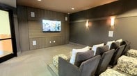 This purpose-built screening room delivers a refined home theater experience through a carefully considered design. The focal point is a large flat-panel display mounted on a feature wall clad in warm wood paneling, which adds texture and visual depth to the space. Built-in speaker cutouts are integrated flush into the paneling, maintaining a clean, uncluttered aesthetic while supporting a full audio-visual setup.

The remaining walls are finished in a deep charcoal tone, promoting contrast and reducing light reflection for optimal viewing conditions. A wall-mounted sconce provides soft, ambient lighting that complements the room's moody, cinema-inspired atmosphere. Recessed ceiling lights offer additional illumination when needed.

Seating consists of generously sized, charcoal upholstered lounge chairs arranged to face the screen, accented with grid-patterned throw pillows. Stone-textured ottomans positioned in front of the seating add a tactile, organic contrast to the otherwise sleek interior. The flooring features low-pile carpet in a neutral tone, contributing to acoustic dampening and overall comfort. The space is compact yet thoughtfully arranged to maximize the immersive viewing experience.
