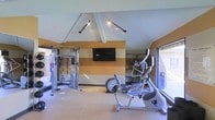 This fitness center offers a thoughtfully designed workout environment that caters to a variety of exercise needs. The space features a vaulted ceiling with angled rooflines that add architectural character and a sense of openness, complemented by track lighting that ensures bright, even illumination throughout. The accent wall showcases a bold striped design in warm orange and neutral tones, adding a vibrant visual focal point anchored by a wall-mounted flat-screen television — ideal for entertainment during workouts. The facility is outfitted with a range of equipment, including a multi-function cable and strength training machine on one side, along with cardio equipment such as an elliptical trainer and a stationary bike. Additional equipment storage or weight stations are visible along the far wall, providing a comprehensive workout setup. Large windows along the perimeter allow natural light to fill the space, creating an inviting and energizing atmosphere. The light-toned flooring is clean and durable, well-suited for a fitness environment. Overall, the space strikes a balance between functional design and aesthetic appeal, making it a practical and motivating place to exercise.