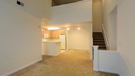 This two-floor space is designed with an open-concept layout on the main level, seamlessly connecting the kitchen and living area. The kitchen features light wood cabinetry, white appliances including a refrigerator and microwave, and a peninsula counter that provides both workspace and a natural divider between the kitchen and the adjoining living area. Track lighting in the kitchen and recessed lighting throughout the main level ensure well-distributed illumination.

The living area is finished with neutral wall-to-wall carpeting that extends throughout the main floor, contributing to a warm and cohesive feel. A striking architectural feature is the soaring double-height ceiling that rises above the main level, creating an expansive, airy atmosphere and enhancing the sense of openness.

A carpeted staircase with a white newel post and railing is positioned to the right, providing access to the upper floor. The clean, neutral color palette of warm beige walls and light-toned finishes throughout lends the space a bright, inviting ambiance. The overall design balances functionality and openness, making effective use of vertical space.