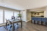 Located at 16202 El Camino Real in Houston, TX, this single-floor space offers a thoughtfully designed open-concept layout that balances comfort and functionality. The main living area makes an immediate impression with its rich hardwood flooring, recessed lighting, and a bold teal accent wall that anchors the room with a contemporary flair. A large sectional sofa, a patterned area rug, and a wall-mounted flat-screen TV create an inviting lounge atmosphere, while a striking brick fireplace with a full-length surround serves as a standout architectural focal point. Decorative wall-mounted accents and a unique live-edge coffee table add artistic character throughout. Natural light flows generously into the space through large windows and sliding glass doors, providing views of the surrounding outdoor area. Adjacent to the main lounge, a dedicated kitchenette features dark granite countertops, white cabinetry, a sink, and a microwave, complemented by an L-shaped breakfast bar with upholstered bar stools — ideal for casual gatherings. A separate workspace area with ample desk space and natural light from nearby windows rounds out the layout, making this space well-suited for both social and professional use.