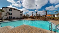 Located at 450 Jimmy Ann Dr in Daytona Beach, Florida, this outdoor amenity space is designed to deliver a resort-style experience under the sunny Florida sky. At its centerpiece is a generously sized swimming pool with crystal-clear turquoise water, framed by a wide, well-appointed paver deck. The surrounding deck offers ample room for lounging, with rows of chaise lounge chairs and shaded umbrella stations positioned to take full advantage of the warm climate and open views of the surrounding landscape, including tall palms and lush greenery on the horizon.

Adjacent to the pool, a covered cabana area provides a refined retreat for relaxation. Each cabana is outfitted with flowing white privacy curtains, comfortable outdoor seating, a low wooden coffee table, and vibrant accent rugs in blue and orange tones, creating a stylish and inviting atmosphere. Ceiling fans overhead ensure comfort even on the warmest days.

A dedicated fitness center is conveniently accessible directly from the pool courtyard, its entrance clearly visible along the paver walkway that connects the various amenity zones. The building architecture features a clean, modern aesthetic with white board-and-batten siding and dark-trimmed windows. The entire outdoor space is enclosed by decorative fencing, offering both security and an open, airy feel throughout.