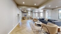 Located at 1905 Concord Blvd in Concord, CA, this thoughtfully designed single-floor space offers a refined blend of contemporary style and functional layout across two distinct areas: a living room and a dining room. Striking herringbone-patterned hardwood flooring runs throughout, providing a warm and visually dynamic foundation that anchors the entire space. The living room is open and inviting, featuring coffered ceiling details, recessed lighting, and a glass-railed overlook accented by elegant geometric pendant fixtures. Arched windows allow natural light to pour in, offering views of the surrounding area including nearby palm trees. The seating arrangement is centered around a marble-topped coffee table, complemented by a neutral-toned area rug that adds texture and depth. A decorative accent wall with botanical-patterned wallpaper adds a sophisticated touch at the far end of the room. The dining room is an intimate yet refined space, highlighted by a bold textured blue accent wall that serves as a dramatic backdrop to a round marble-top dining table paired with upholstered mid-century style chairs. White louvered double doors and a frosted glass entry door add architectural character. Together, these spaces create a cohesive, elegantly curated environment ideal for both relaxation and social gatherings.