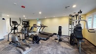 This fitness center is a single-floor facility thoughtfully designed to accommodate a wide range of exercise needs. The space is outfitted with modern, patterned carpet flooring and illuminated by recessed ceiling lighting, creating a clean and energizing environment. The walls are finished in a soft, neutral yellow-green tone, contributing to a calm and motivating atmosphere.

The room is furnished with a solid lineup of cardio equipment, including treadmills, an elliptical trainer, and a stationary bike, all from the SportFit brand. These machines are arranged to allow comfortable spacing and ease of movement between stations. A window positioned along the far wall allows natural light to enter the space, enhancing the overall workout environment.

On the strength-training side, the facility includes a multi-station cable and weight machine system, complete with a weight stack and illustrated exercise guides mounted directly on the unit, making it accessible for users of all experience levels. Additional strength equipment is visible toward the back of the room, offering further versatility.

Practical amenities include ceiling-mounted ventilation, a smoke detector, a security camera, and a clearly marked emergency exit. Overall, this fitness center is a functional and well-maintained space built to support both cardio and resistance training.