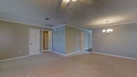This single-floor space presents a generously sized open room with soft, neutral-toned carpeting that extends throughout, creating a warm and cohesive foundation. The walls are painted in a calming light gray-blue hue, complemented by crisp white crown molding that runs along the ceiling perimeter, adding a refined architectural detail. The ceiling features a textured finish and is equipped with a ceiling fan as well as a chandelier-style light fixture, providing both functional and decorative lighting options. Multiple doorways are visible throughout the space, leading to what appear to be adjoining rooms and hallways, suggesting a well-connected floor plan. White-paneled doors and bright white baseboards provide clean contrast against the soft wall color. HVAC vents are positioned thoughtfully in both the ceiling and walls, ensuring efficient climate control. The overall ambiance of the space is bright, open, and neutral, making it highly versatile and easy to personalize. The combination of classic molding details, consistent flooring, and a generous footprint gives the room a comfortable and functional character suited to a variety of uses.