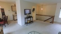 This single-floor space showcases an open, flowing layout highlighted by neutral tones and plush carpeting throughout the main living area. A central structural column serves as a natural divider, anchoring a wall-mounted flat-screen television above a dark wood console table styled with decorative accents. The open design allows sightlines across multiple zones, creating a sense of spaciousness.

Adjacent to the living area, a kitchen is partially visible, featuring light-toned cabinetry and white appliances, offering a functional workspace that integrates seamlessly with the surrounding open floor plan. A staircase with vertical wood balusters lines one edge of the space, contributing an architectural detail that adds visual structure to the layout.

Overhead, a pendant light with a warm, stained-glass-style shade hangs in a secondary area, casting a soft ambient glow and adding a touch of character to the otherwise clean, understated interior. The overall ambiance is calm and practical, with a neutral color palette throughout that lends itself well to a variety of furnishing and décor styles.