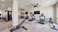 Located in Conroe, Texas, this fitness center offers a comprehensive and thoughtfully designed exercise environment spread across a single, open floor. The space is outfitted with a wide variety of cardio equipment, including multiple treadmills, stair climbers, and elliptical machines, arranged to allow comfortable circulation throughout the room. A dedicated strength training area features a cable machine, a power rack with weight plates, a full dumbbell rack with tiered weight options, and adjustable benches, catering to a broad range of workout routines. Wall-mounted flat-screen televisions are positioned for visibility across the floor, while large, multi-blade ceiling fans promote consistent air circulation throughout the space. The interior is finished with neutral-toned carpet tile flooring and crisp white walls, contributing to a clean, modern aesthetic. Floor-to-ceiling mirrors along one wall enhance the sense of openness and allow for form monitoring during strength exercises. Natural light filters in through large windows and glass doors that face an outdoor area, keeping the environment bright and inviting. Recessed lighting supplements the natural light for consistent illumination. Structural columns are integrated into the layout without disrupting the overall flow of the space, and restroom access is conveniently available within the facility.