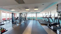 This well-appointed fitness center at 1000 S Broadway in Denver, Colorado, offers a comprehensive workout environment thoughtfully designed across a single floor. The main gym area is bright and spacious, featuring large floor-to-ceiling windows that flood the space with natural light while framing sweeping views of the Denver skyline and surrounding landscape. Teal accent walls and recessed panel lighting add a modern, energetic aesthetic throughout.

The cardio zone is equipped with a row of treadmills and elliptical machines, all positioned to take advantage of the panoramic views. Strength training is well-represented with a variety of cable machines, weight benches, dumbbells, and kettlebells, neatly organized along mirrored walls that enhance the sense of space. Durable rubber flooring with striped detailing covers the main workout area, providing both safety and a polished look.

Adjacent to the main gym is a dedicated studio room featuring rich wood-look flooring, full-length mirrors, a ballet barre, a wall-mounted flat-screen TV, and spin bikes, making it ideal for group classes or personal training. The entry area showcases a striking textured accent wall and glass doors that connect seamlessly to the gym. A tiled lobby corridor with carpet tile flooring and a tanning booth rounds out the facility's amenities, offering a complete wellness destination.