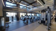 This contemporary fitness center offers a thoughtfully designed workout environment that combines functionality with an open, energizing atmosphere. The space is defined by its striking soaring ceilings, featuring a bold coffered ceiling structure with integrated linear lighting that provides consistent, well-distributed illumination throughout. Large floor-to-ceiling windows line the perimeter walls, flooding the interior with abundant natural light and offering views of the surrounding outdoors, creating an inviting and motivating setting for exercise. The flooring transitions between durable dark rubber gym matting in the primary workout zones and lighter-toned flooring in adjacent areas, providing both safety and visual contrast. The facility is outfitted with a robust selection of cardio equipment, including multiple treadmills and elliptical machines, all arranged in organized rows to maximize usability and flow. Complementing the cardio section, a variety of strength training machines and cable systems are positioned throughout the space, catering to a full range of fitness goals. The combination of modern architectural design, premium equipment, and thoughtful layout makes this fitness center a well-rounded, high-quality facility suited for diverse exercise routines.