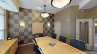 This contemporary building lobby is designed with a polished, modern aesthetic that makes a strong first impression. The main entrance features large windows that flood the space with natural light, offering views of the surrounding streetscape. The entry area transitions seamlessly between rich stone-look tile flooring and plush carpeting, defined by sweeping curved architectural ceiling elements in warm golden and deep brown tones that add visual dynamism to the space.

A comfortable seating area near the entrance includes modern sofas and armchairs arranged around a bold red accent table, providing a welcoming lounge for residents and guests. Adjacent to the lobby, a dedicated mailroom alcove houses an extensive bank of individual mailboxes, conveniently located near a pair of stainless steel elevator doors framed by dark tile surrounds.

The corridors extending from the lobby are lined with dark wood paneling, recessed lighting, and curved walls that guide the eye through the building with architectural elegance. A fitness room is accessible directly from the hallway, visible through a glazed door.

The conference room is a standout feature, showcasing a striking checkerboard accent wall in alternating gold and charcoal tones. A light wood conference table is surrounded by black leather chairs beneath a pendant light fixture, creating a professional yet inviting meeting environment. Built-in cabinetry completes the room's functional design.