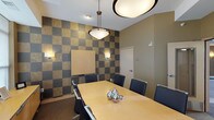 This contemporary building lobby is designed with a polished, modern aesthetic that makes a strong first impression. The main entrance features large windows that flood the space with natural light, offering views of the surrounding streetscape. The entry area transitions seamlessly between rich stone-look tile flooring and plush carpeting, defined by sweeping curved architectural ceiling elements in warm golden and deep brown tones that add visual dynamism to the space.

A comfortable seating area near the entrance includes modern sofas and armchairs arranged around a bold red accent table, providing a welcoming lounge for residents and guests. Adjacent to the lobby, a dedicated mailroom alcove houses an extensive bank of individual mailboxes, conveniently located near a pair of stainless steel elevator doors framed by dark tile surrounds.

The corridors extending from the lobby are lined with dark wood paneling, recessed lighting, and curved walls that guide the eye through the building with architectural elegance. A fitness room is accessible directly from the hallway, visible through a glazed door.

The conference room is a standout feature, showcasing a striking checkerboard accent wall in alternating gold and charcoal tones. A light wood conference table is surrounded by black leather chairs beneath a pendant light fixture, creating a professional yet inviting meeting environment. Built-in cabinetry completes the room's functional design.