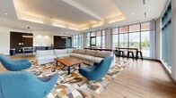 Located at 140 Ferguson Dr in Woodstock, Ontario, this single-floor amenity lounge offers a thoughtfully designed open-concept space that balances comfort, style, and functionality. The living area is generously sized, providing ample room for both relaxation and social activity.

The interior design is defined by a clean, contemporary aesthetic. Striking vertical wood slat accent walls serve as a bold architectural feature, complementing the warm-toned light hardwood flooring that runs throughout. A large flat-screen television is mounted seamlessly into one of the slatted wall panels, creating a polished entertainment focal point. The coffered ceiling with integrated recessed and cove lighting adds dimension and a refined ambiance to the space.

Floor-to-ceiling windows along multiple walls flood the interior with abundant natural light and offer open views of the surrounding greenery and landscaped exterior — including a stone retaining wall and outdoor gathering areas visible from within. The windows also provide direct access to an outdoor patio, blurring the boundary between indoor and outdoor spaces.

The lounge is furnished with plush sectional sofas, sculptural blue accent chairs, and a solid wood coffee table, all arranged over a geometric patterned area rug. A ping-pong table and bar-height seating along the windows round out the recreational offerings, making this a versatile and inviting communal retreat.