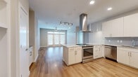 This thoughtfully designed 2-bedroom, 2-bathroom residence offers a seamless blend of modern aesthetics and functional living across a single floor. The open-concept layout connects the kitchen and living room, creating a bright, airy atmosphere enhanced by light wood-look flooring throughout the main living areas.

The kitchen is a standout feature, equipped with sleek white flat-panel cabinetry, marble-look countertops, a full suite of stainless steel appliances including a range, dishwasher, and French door refrigerator, and a prominent island range hood. The living room is filled with natural light from a set of glass panel doors with transom windows, complemented by a ceiling fan overhead.

The primary bedroom is generously sized, featuring plush carpet flooring, a ceiling fan, and a bright window. It is paired with a well-appointed en-suite bathroom that includes a double-sink vanity with marble-look countertops, wood-look flooring, and a glass-enclosed walk-in shower. Both bedrooms include dedicated walk-in closets with built-in multi-tier shelving and hanging rods for organized storage.

The second bedroom mirrors the comfort of the primary, with carpet flooring, a ceiling fan, and natural light from a large window. The overall design maintains a clean, neutral color palette, contributing to a cohesive and modern ambiance throughout.
