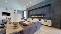 This thoughtfully designed single-floor space combines modern sophistication with warm, rustic touches to create an inviting communal environment. The centerpiece of the main lounge is a striking floor-to-ceiling dark metal tile feature wall housing an expansive linear fireplace, which anchors the room and creates a dramatic focal point. Generous seating — including a large sectional sofa and a built-in upholstered bench — surrounds the fireplace, encouraging relaxed gathering. Vaulted shiplap ceilings and recessed lighting add architectural interest and a sense of openness overhead, while light wood-look flooring flows seamlessly throughout the space.

A dedicated kitchenette area is outfitted with a central island, wall-mounted television, open wooden shelving, under-counter storage, and industrial-style pendant lighting — offering both functionality and style. A dining area with a solid wood table and mixed seating is bathed in natural light from large windows and French doors that open directly to an outdoor patio. Rustic sliding barn doors with vertical metal slats connect the lounge to a welcoming entry corridor, adding a distinctive architectural detail. Throughout, muted blue-grey walls and natural wood accents create a cohesive, contemporary-rustic ambiance.
