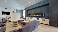 This thoughtfully designed single-floor space combines modern sophistication with warm, rustic touches to create an inviting communal environment. The centerpiece of the main lounge is a striking floor-to-ceiling dark metal tile feature wall housing an expansive linear fireplace, which anchors the room and creates a dramatic focal point. Generous seating — including a large sectional sofa and a built-in upholstered bench — surrounds the fireplace, encouraging relaxed gathering. Vaulted shiplap ceilings and recessed lighting add architectural interest and a sense of openness overhead, while light wood-look flooring flows seamlessly throughout the space.

A dedicated kitchenette area is outfitted with a central island, wall-mounted television, open wooden shelving, under-counter storage, and industrial-style pendant lighting — offering both functionality and style. A dining area with a solid wood table and mixed seating is bathed in natural light from large windows and French doors that open directly to an outdoor patio. Rustic sliding barn doors with vertical metal slats connect the lounge to a welcoming entry corridor, adding a distinctive architectural detail. Throughout, muted blue-grey walls and natural wood accents create a cohesive, contemporary-rustic ambiance.