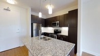 Located at 1005 West 5th Avenue in Columbus, Ohio's vibrant Grandview Heights neighborhood, this single-floor residence offers a thoughtfully designed living environment with contemporary finishes throughout. The kitchen stands as a central highlight, showcasing rich, dark espresso cabinetry that extends to the ceiling for generous storage capacity. Gleaming granite countertops provide ample prep space, while a large kitchen island with an integrated sink serves as both a functional workspace and a natural gathering point. Stainless steel appliances — including a refrigerator, electric range, built-in microwave, and dishwasher — complete the modern culinary setup. Elegant pendant lighting hangs above the island, casting a warm, focused glow over the space. A classic white tile backsplash adds a clean contrast to the dark cabinetry, enhancing the kitchen's polished aesthetic. The flooring transitions seamlessly from the kitchen into the adjoining living areas, contributing to the open, cohesive flow of the layout. The overall ambiance is modern and refined, combining practical design with stylish details to create a comfortable and visually appealing living space.