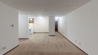 Located at 825 9th Ave S in Saint Cloud, Minnesota, this single-floor unit offers a practical and comfortable layout designed for everyday living. The open living area is generously proportioned, featuring warm beige carpeting, neutral white walls, and a textured ceiling that adds subtle character throughout. A large sliding window draws in natural light and provides a pleasant view of the surrounding greenery, creating a bright and welcoming atmosphere. Baseboard heating lines the perimeter, ensuring comfort throughout the colder Minnesota seasons.

The kitchen is efficiently arranged with dark wood cabinetry, a double-basin sink with laminate countertops, tile flooring, and white appliances including a refrigerator and electric range. The adjacent layout allows for easy flow between the kitchen and living spaces. A structural column defines the transition between the kitchen and living areas, adding an architectural element to the open floor plan.

The bedroom is carpeted and well-lit, featuring a window with an outdoor view and a spacious closet equipped with a full-length hanging rod and upper shelving, offering ample storage. The bathroom includes a single-sink vanity with dark wood cabinetry, a tub-shower combination, mosaic tile flooring, and a ceiling vent. The dark wood door frames and trim throughout the unit provide a cohesive, classic finish.