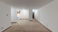 Located at 825 9th Ave S in Saint Cloud, Minnesota, this single-floor unit offers a practical and comfortable layout designed for everyday living. The open living area is generously proportioned, featuring warm beige carpeting, neutral white walls, and a textured ceiling that adds subtle character throughout. A large sliding window draws in natural light and provides a pleasant view of the surrounding greenery, creating a bright and welcoming atmosphere. Baseboard heating lines the perimeter, ensuring comfort throughout the colder Minnesota seasons.

The kitchen is efficiently arranged with dark wood cabinetry, a double-basin sink with laminate countertops, tile flooring, and white appliances including a refrigerator and electric range. The adjacent layout allows for easy flow between the kitchen and living spaces. A structural column defines the transition between the kitchen and living areas, adding an architectural element to the open floor plan.

The bedroom is carpeted and well-lit, featuring a window with an outdoor view and a spacious closet equipped with a full-length hanging rod and upper shelving, offering ample storage. The bathroom includes a single-sink vanity with dark wood cabinetry, a tub-shower combination, mosaic tile flooring, and a ceiling vent. The dark wood door frames and trim throughout the unit provide a cohesive, classic finish.