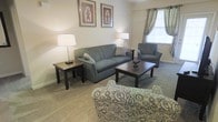 This single-floor space offers a comfortable and thoughtfully arranged living environment. The living room serves as the centerpiece of the space, featuring a plush sofa and coordinating armchair in a muted blue-gray tone, creating a cohesive and relaxing atmosphere. A rectangular coffee table and matching end tables in a rich dark wood finish provide both functionality and visual contrast against the soft carpeted flooring. Ambient lighting is provided by a pair of classic table lamps positioned on either side of the seating area, casting a warm and inviting glow throughout the room. Natural light filters in through a curtained window dressed with elegant patterned drapes, adding texture and softness to the overall aesthetic. The walls are kept in a neutral, light tone, allowing the framed decorative artwork above the sofa to serve as a subtle focal point. The overall ambiance of the space is calm and residential, with a traditional design sensibility that emphasizes comfort and practicality. The layout makes efficient use of the available floor space, with furniture arranged to encourage conversation and relaxation. The carpeted flooring adds warmth underfoot, contributing to the cozy, lived-in feel of this welcoming single-level residence.