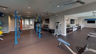 Located at 9511 Wes Kearney Way in Riverview, Florida, this fitness center is designed to support a comprehensive range of workout routines within a thoughtfully organized, single-floor layout. The facility is divided into distinct functional zones that flow seamlessly into one another, accommodating both cardio and strength training needs.

The cardio zone is lined with rows of treadmills, ellipticals, stationary bikes, and rowing machines positioned along a bold navy blue accent wall. Large windows with horizontal blinds line this section, allowing abundant natural light to fill the space while offering views of the surrounding community. Ceiling fans overhead provide additional air circulation throughout.

An adjacent strength training area features a blue cable and functional training rig, a dumbbell rack, weight benches, and a punching bag, providing ample options for resistance and functional fitness work. Wall-mounted flat-screen televisions are positioned throughout for entertainment during workouts. The flooring throughout consists of dark rubberized material suited for heavy equipment and high-traffic use.

A utility counter with open shelving runs along one wall, offering space for amenities and storage. The overall color palette — grey walls, navy accents, and white trim — gives the facility a clean, modern aesthetic that is both energizing and functional.