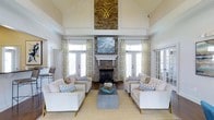 This sophisticated single-floor space makes an immediate impression with its soaring vaulted ceilings and airy, open atmosphere. The focal point of the main living area is a striking floor-to-ceiling stacked stone fireplace, crowned with a decorative gold emblem and complemented by a crisp white mantel. A large piece of wall art is mounted above the mantel, adding a refined artistic touch to the ensemble.

The seating arrangement is thoughtfully curated, featuring neutral-toned sofas and armchairs accented with blue and white geometric throw pillows, all centered around a sleek rectangular coffee table. A natural fiber area rug anchors the space, adding warmth and texture to the light hardwood flooring beneath.

Natural light floods the room through multiple sets of French doors and tall windows, each dressed with elegant patterned drapery in soft gold and ivory tones. The doors offer what appears to be access to outdoor areas, enhancing the sense of openness and connection to the surrounding environment.

The overall ambiance is bright, welcoming, and polished — a harmonious blend of coastal-inspired accents and classic architectural detailing. The high ceilings, abundant glazing, and carefully selected furnishings combine to create a space that feels both grand in scale and comfortably inviting.
