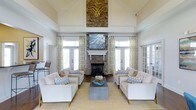 This sophisticated single-floor space makes an immediate impression with its soaring vaulted ceilings and airy, open atmosphere. The focal point of the main living area is a striking floor-to-ceiling stacked stone fireplace, crowned with a decorative gold emblem and complemented by a crisp white mantel. A large piece of wall art is mounted above the mantel, adding a refined artistic touch to the ensemble.

The seating arrangement is thoughtfully curated, featuring neutral-toned sofas and armchairs accented with blue and white geometric throw pillows, all centered around a sleek rectangular coffee table. A natural fiber area rug anchors the space, adding warmth and texture to the light hardwood flooring beneath.

Natural light floods the room through multiple sets of French doors and tall windows, each dressed with elegant patterned drapery in soft gold and ivory tones. The doors offer what appears to be access to outdoor areas, enhancing the sense of openness and connection to the surrounding environment.

The overall ambiance is bright, welcoming, and polished — a harmonious blend of coastal-inspired accents and classic architectural detailing. The high ceilings, abundant glazing, and carefully selected furnishings combine to create a space that feels both grand in scale and comfortably inviting.