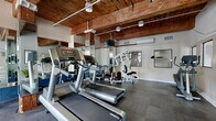 This fitness facility offers a thoughtfully designed exercise space that blends rustic architectural character with modern gym functionality. The room's most distinctive design feature is its exposed wooden ceiling beams, which lend a warm, industrial-rustic ambiance to the space. White walls and large windows allow natural light to filter in, creating a bright and inviting atmosphere. Dark hardwood-style flooring runs throughout, providing both durability and a clean, polished look.

The exercise room is comprehensively outfitted with a variety of cardio equipment, including multiple treadmills, an elliptical trainer, a stationary bike, and a stair stepper. A multi-functional cable weight machine and a dedicated bench press station address strength training needs. Full-length mirrors line portions of the walls, visually expanding the space and allowing users to monitor their form during workouts. A wall-mounted flat-screen television adds convenience for entertainment during exercise sessions.

A split-unit air conditioning system ensures climate control throughout the year. Additional amenities include package lockers along one wall, potted plants for a touch of greenery, and clearly posted gym rules. The space is well-lit with track lighting and offers views of the outdoors through its windows, making for an energizing workout environment.